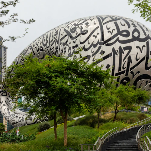 Futuristic building with Arabic calligraphy, surrounded by lush greenery and modern skyscrapers in the background.