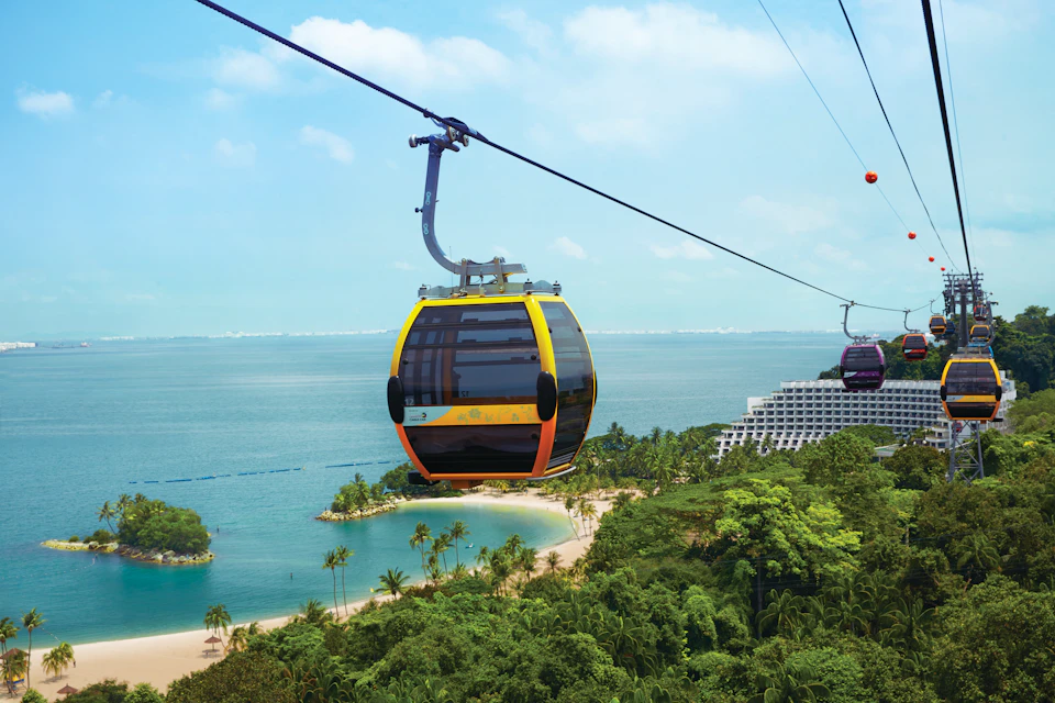 A yellow cable car glides over lush greenery and a sandy beach with ocean views under a partly cloudy sky.
