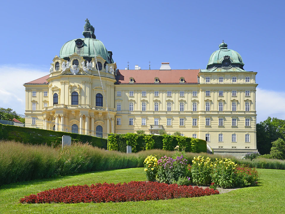 A yellow baroque-style building with green domes, landscaped gardens, and colorful flowerbeds under a clear blue sky.