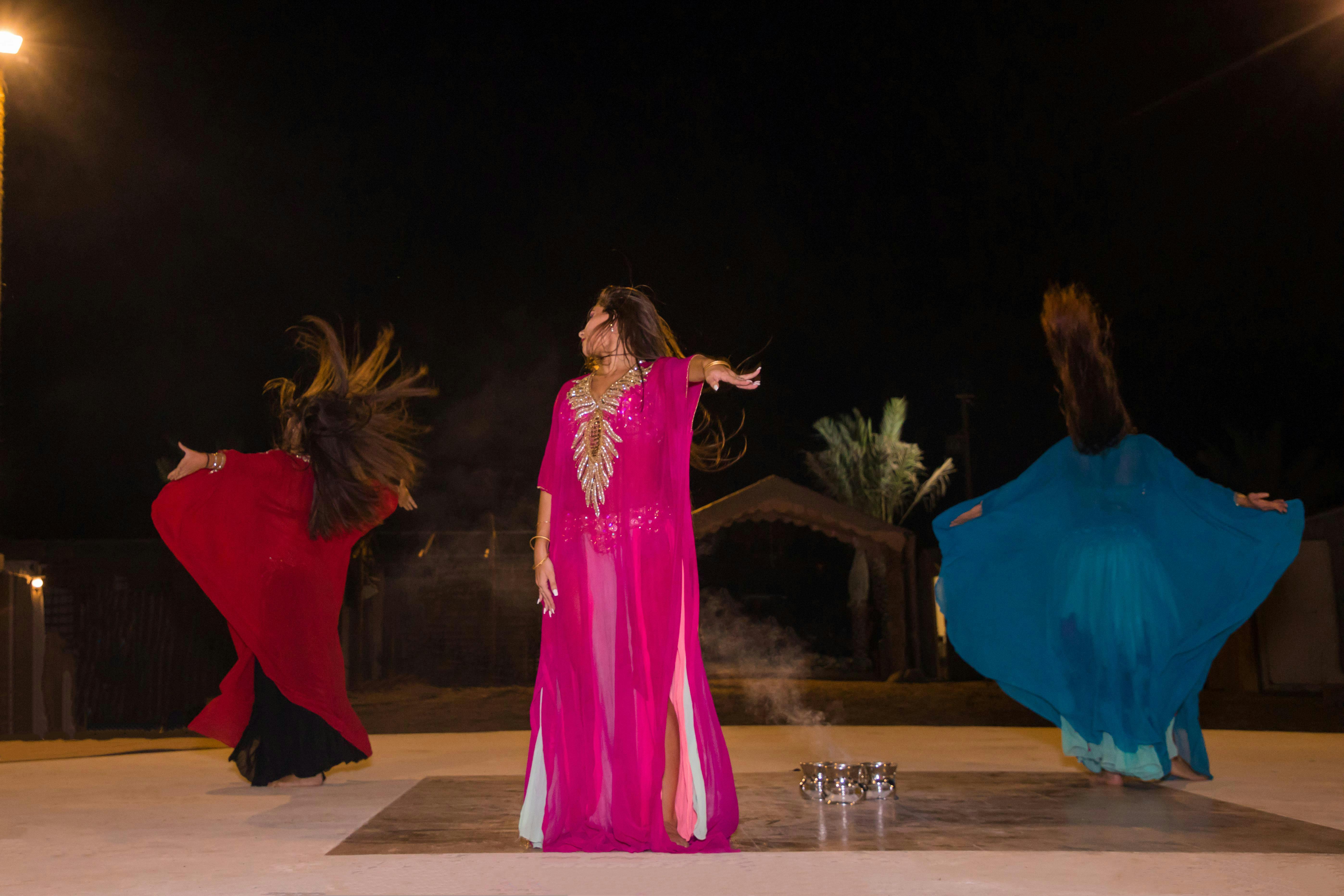 Tre persone in abiti colorati che ballano energicamente su un palco di notte, con le palme sullo sfondo.