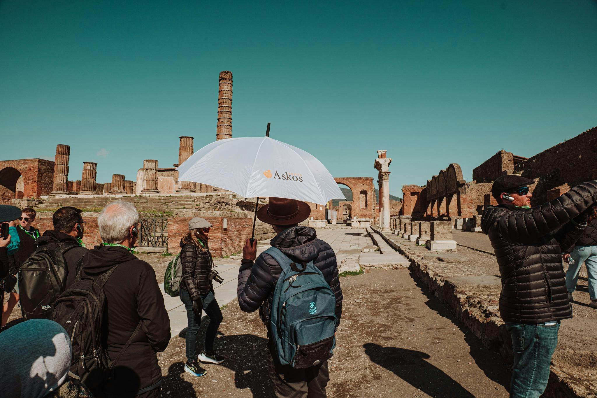 A group of people, some with backpacks, stand near ancient ruins under a clear sky. One person holds a white umbrella marked 'Askos.'