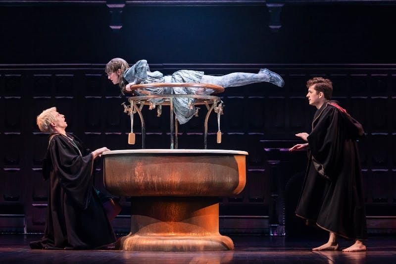 Una persona in vestaglia si trova accanto a una figura fluttuante avvolta in un tessuto, illuminata in modo drammatico su un palcoscenico buio.