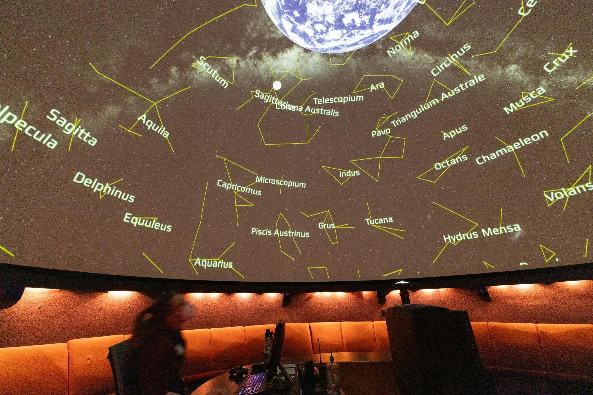 Projekce planetária ukazující souhvězdí a Zemi, s osobou a stoly v popředí.