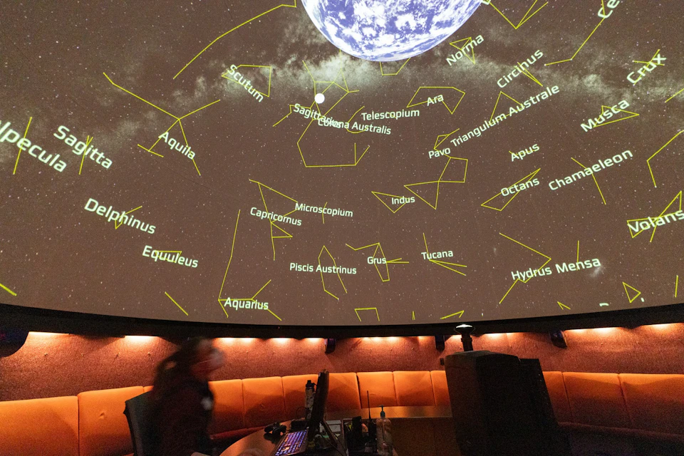 A planetarium projection showing constellations and Earth, with a person and desks in the foreground.