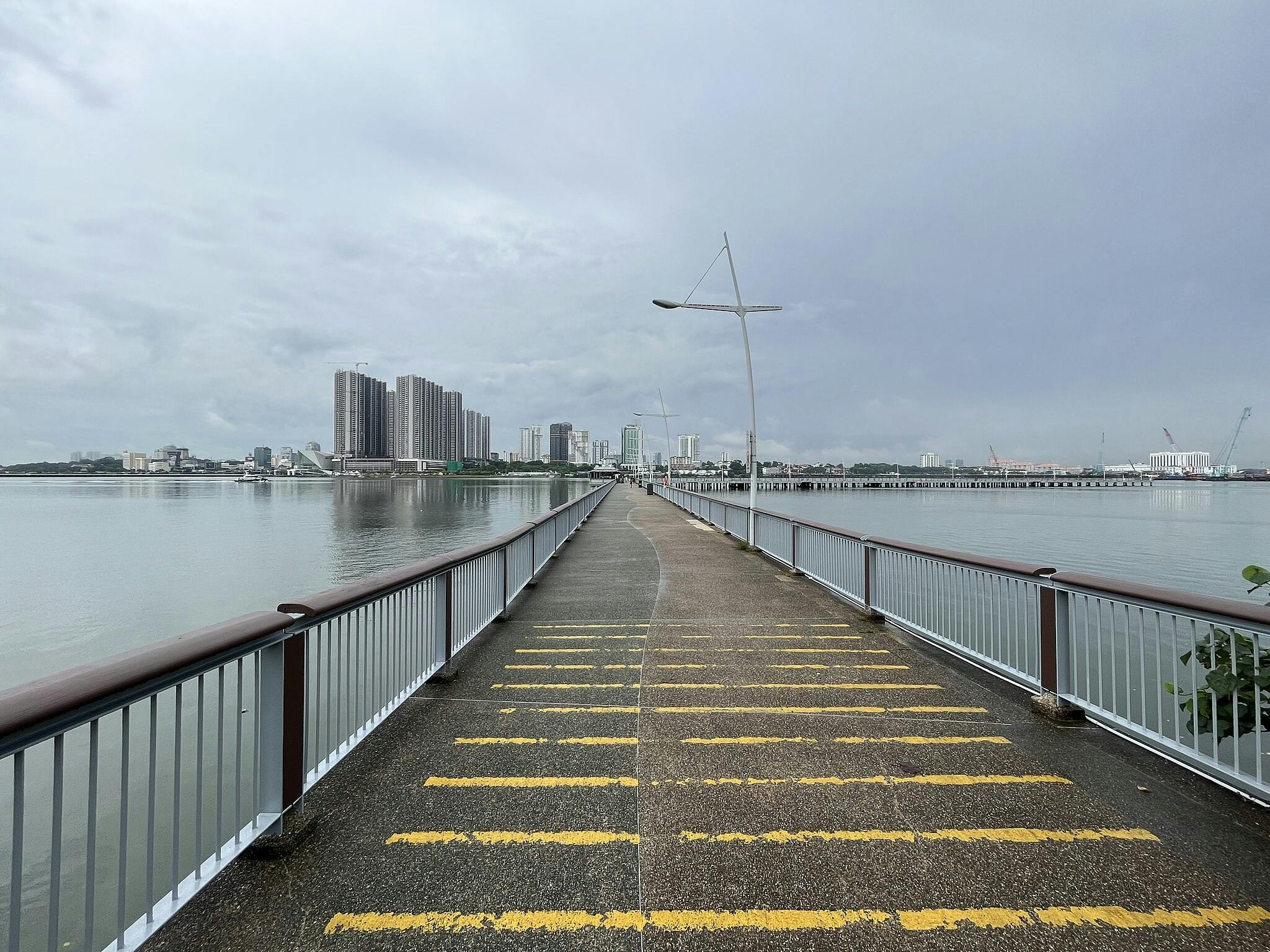 A long pathway with yellow stripes leads to a city skyline beside a body of water, with buildings and cranes in the distance.