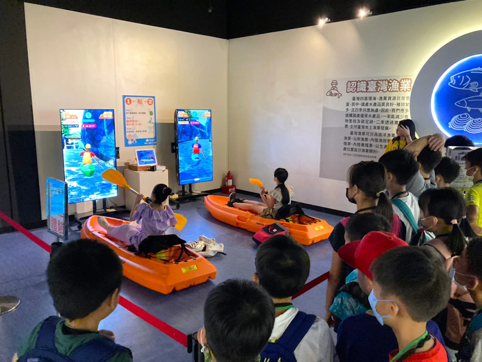 People sitting in kayak simulators in front of screens while a group watches in an indoor space.