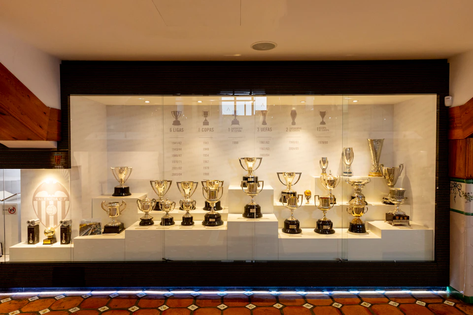A glass display case showcasing multiple sports trophies on shelves with a white background listing years and titles.