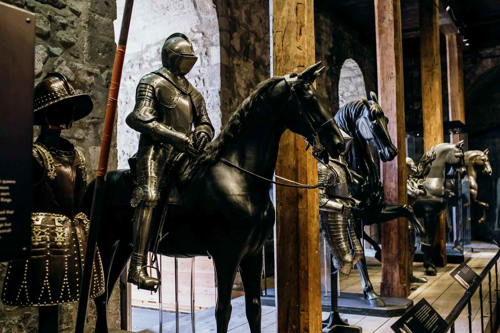 Two armored knight mannequins on horseback in a dimly lit stone-walled room with large arched windows.