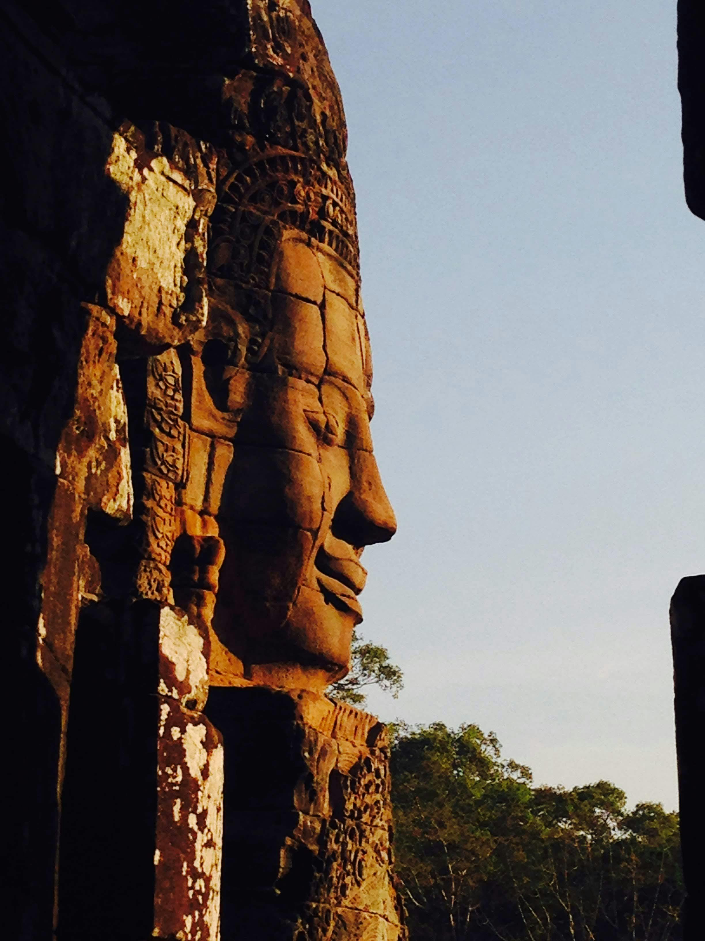 Profile of a stone-carved face on an ancient temple with a clear sky and trees in the background.
