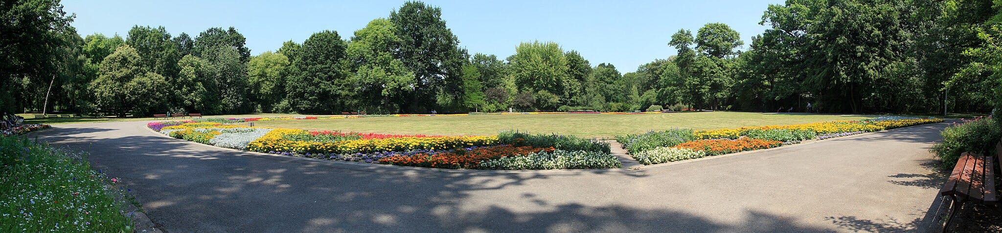 Ένα πάρκο με πολύχρωμα παρτέρια που σχηματίζουν ένα κυκλικό μοτίβο, που περιβάλλεται από ψηλά πράσινα δέντρα και πολλά παγκάκια στα μονοπάτια.