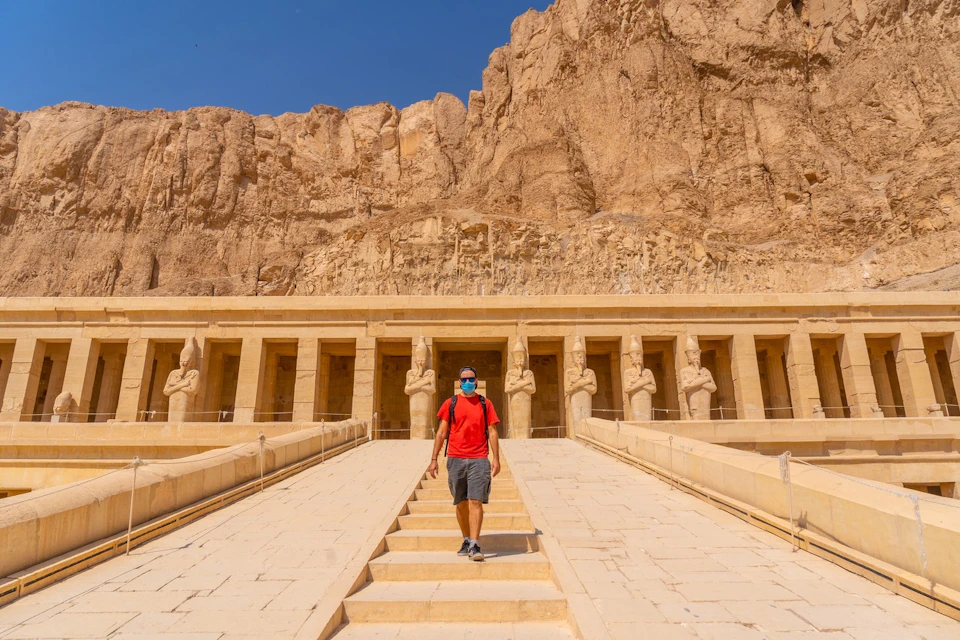 A person in a red shirt walking down stairs in front of an ancient temple carved into a cliff under a clear blue sky.