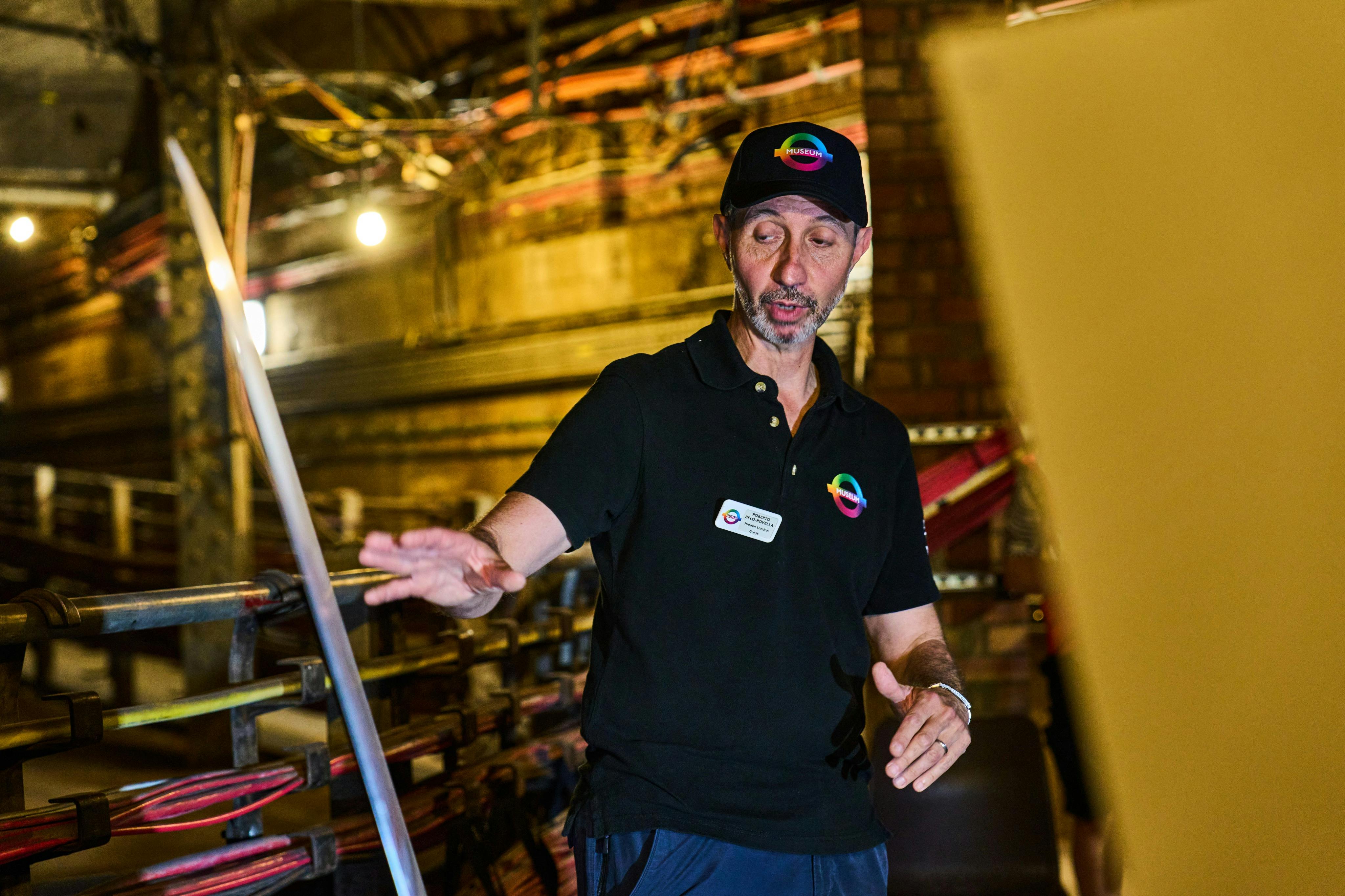 A person wearing a black shirt and cap with a colorful logo gestures in a workshop with tools and equipment in the background.