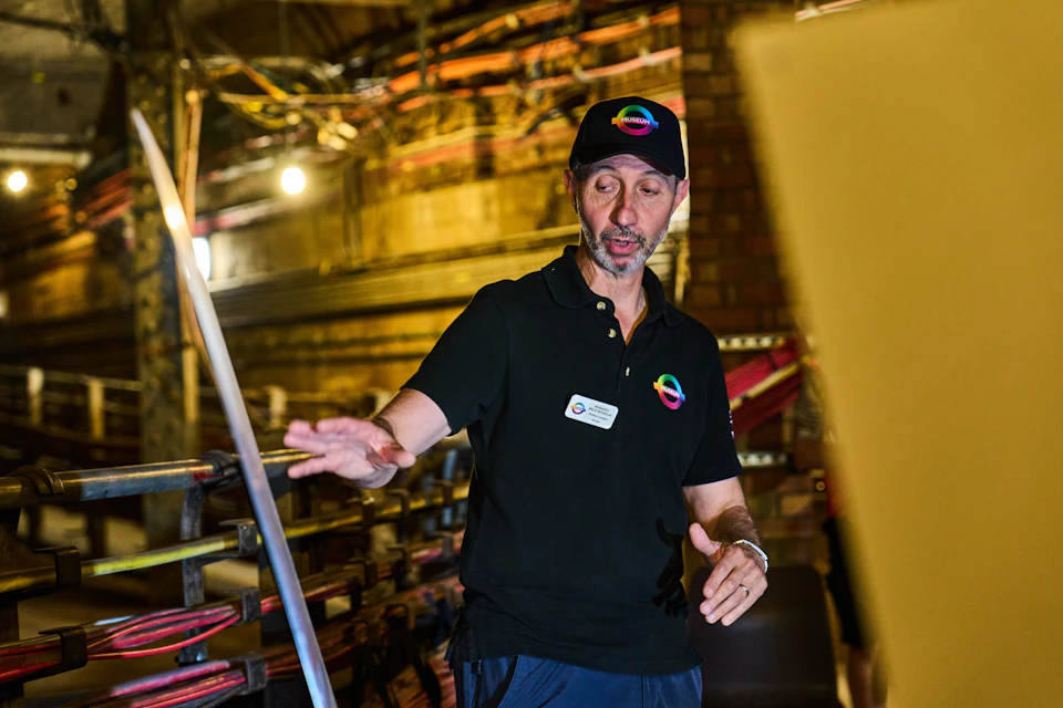 A person wearing a black shirt and cap with a colorful logo gestures in a workshop with tools and equipment in the background.