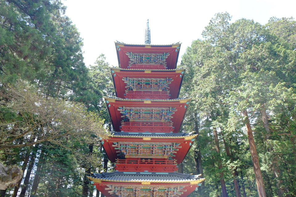 A red multi-tiered pagoda surrounded by tall green trees under a bright sky.