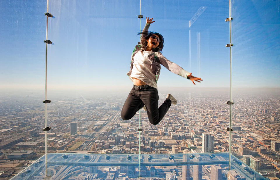 Person jumping with joy in a glass observation box, high above a sprawling cityscape under a clear blue sky.