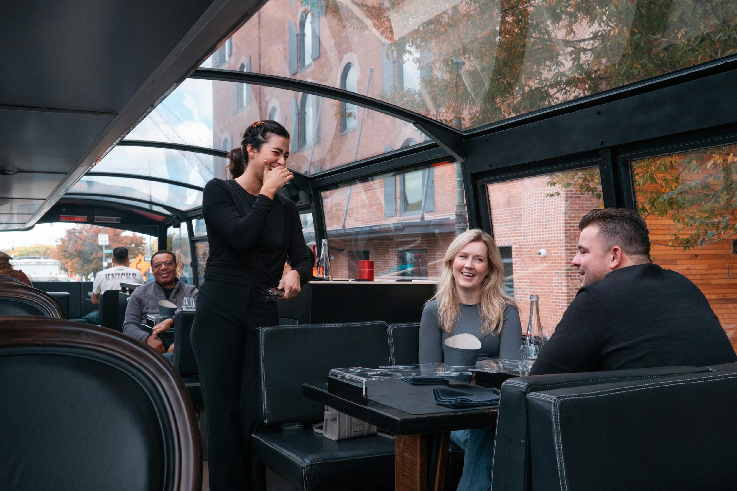 Een vrouw staat glimlachend en haar mond bedekkend in een bus met glazen dak, terwijl passagiers op hun stoel zitten en lachen.