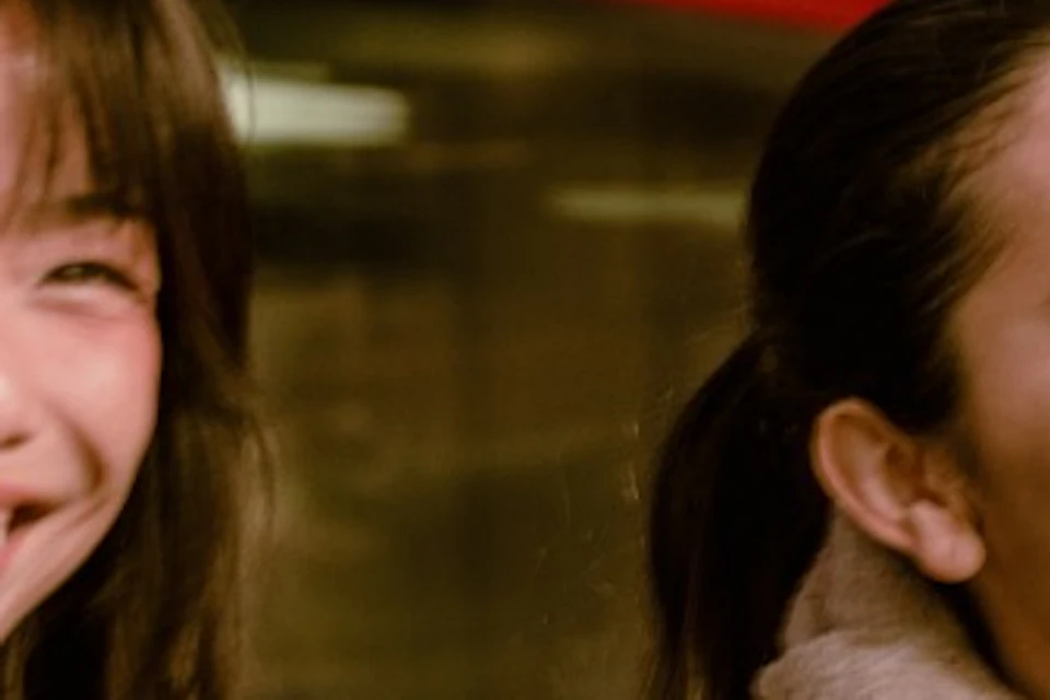 Two women smiling and chatting on a city street with a blurred red double-decker bus passing by in the background.