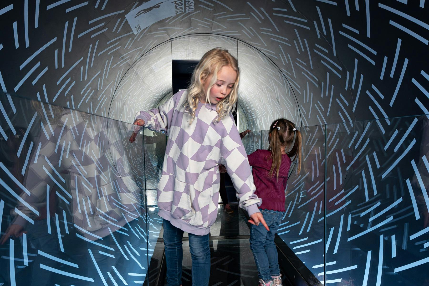 Δύο παιδιά περπατούν μέσα από μια σήραγγα με ανακλαστικούς τοίχους και ένα εσωτερικό με μοτίβα.