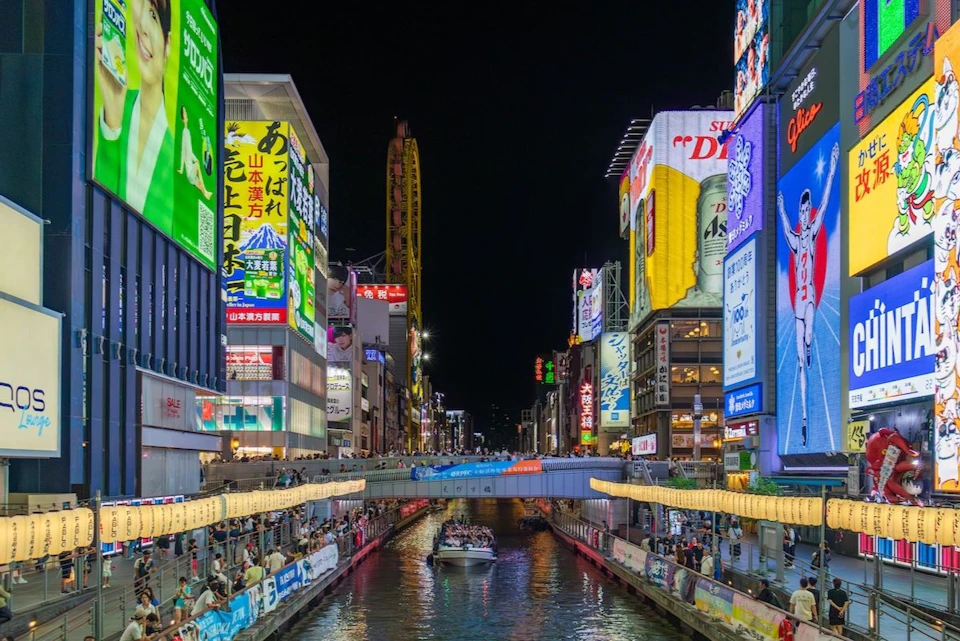 Vibrant cityscape at night showing neon-lit buildings, a canal with a boat, and people walking along both sides of the waterway.