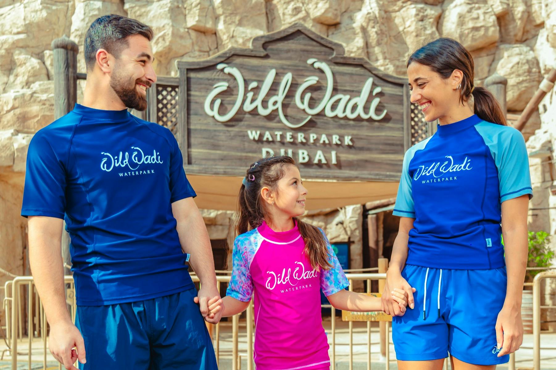 Une famille de trois personnes, portant des chemises du parc aquatique Wild Wadi, se tient devant le panneau d'entrée du parc aquatique Wild Wadi.