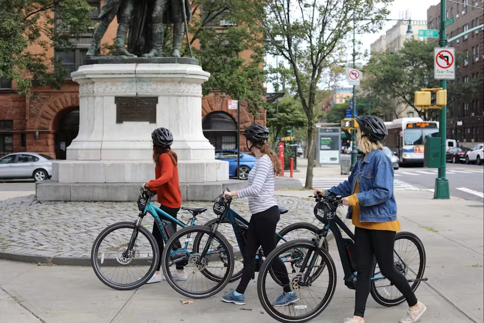 Three people wearing helmets stand beside bicycles near a statue in an urban park setting.