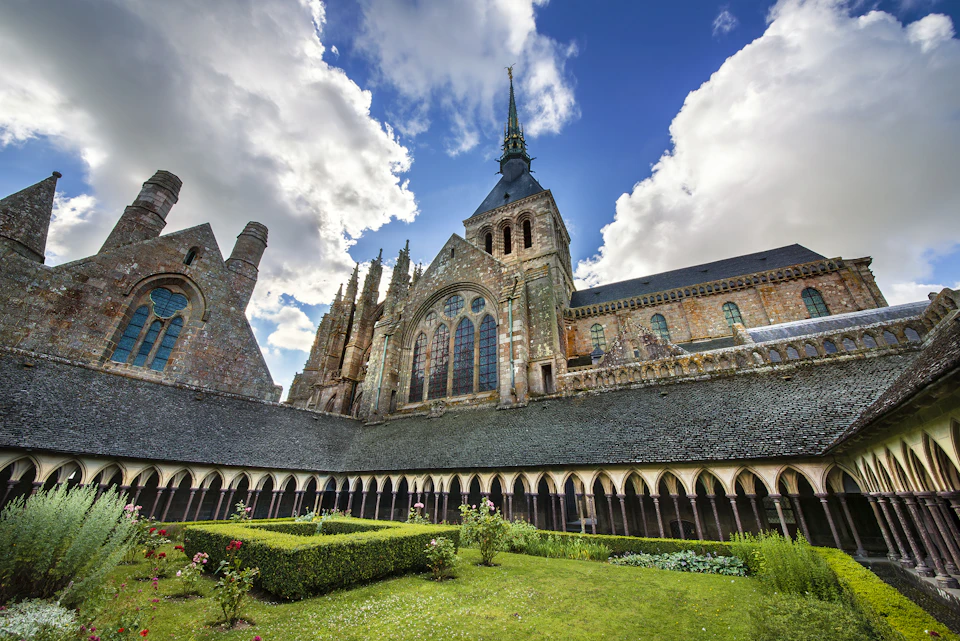 Historic stone abbey with tall spire, arched windows, and a manicured garden courtyard under a partly cloudy sky.
