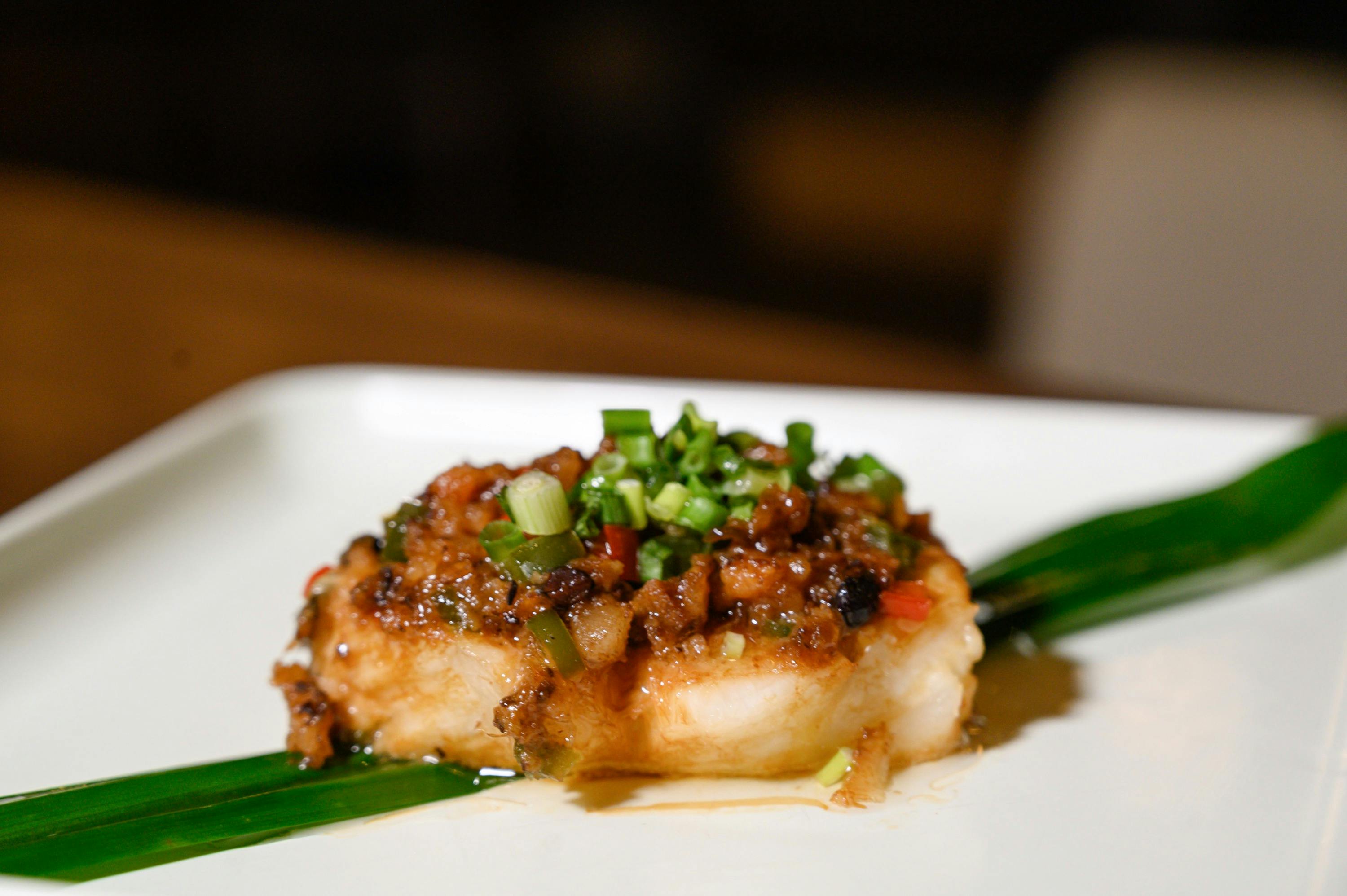 A plated dish featuring a piece of fish topped with a savory sauce and garnished with chopped green onions.