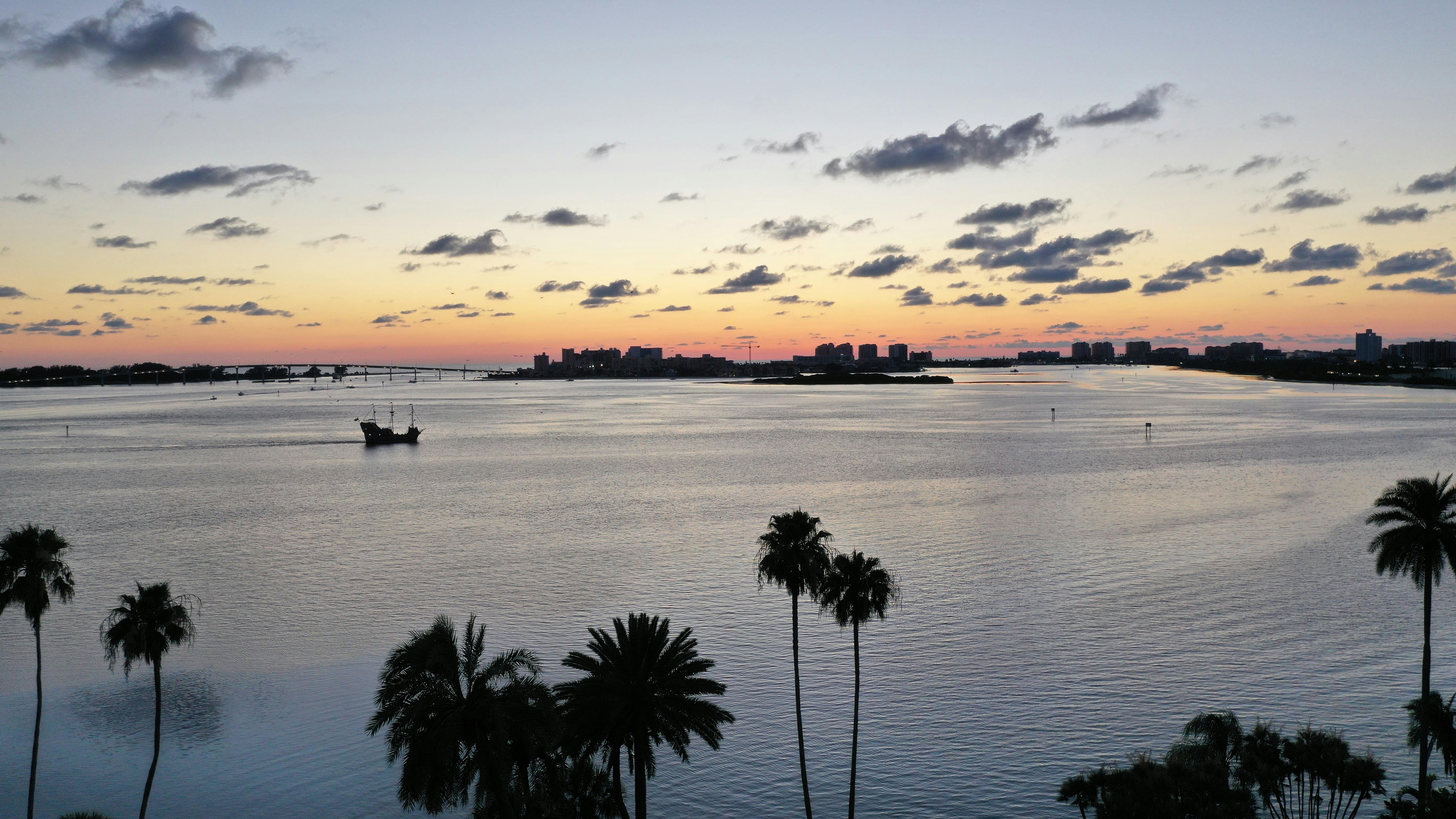 실루엣의 해적선, 야자수, 멀리 떨어진 건물, 흩어져 있는 구름이 있는 잔잔한 만 위로 석양을 감상해보세요.