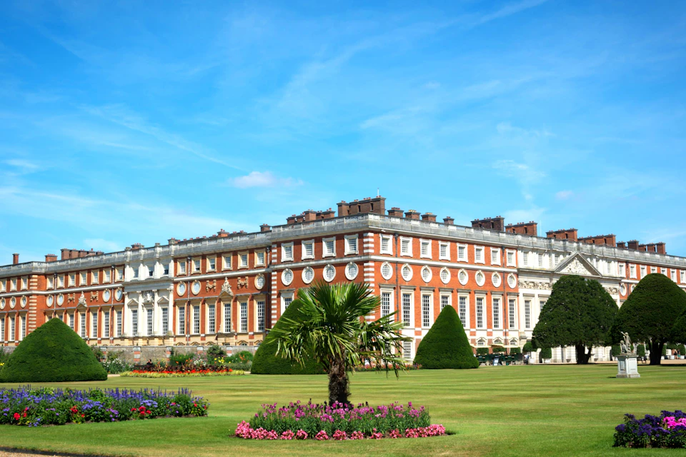 A large, ornate building with red and white exterior sits behind a well-manicured garden with colorful flowers and trimmed hedges.