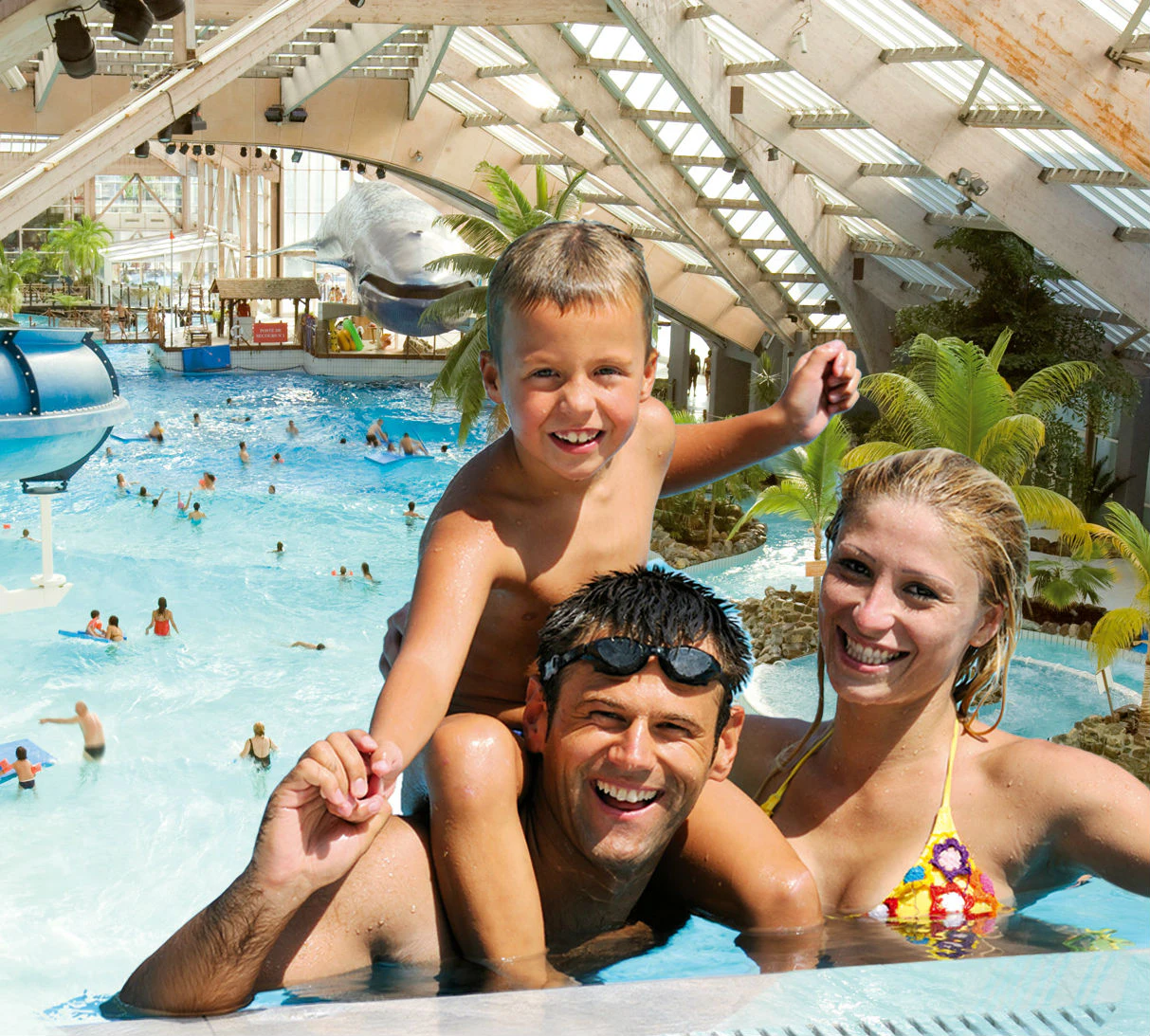 A smiling family of three in a tropical-themed indoor pool with a young boy on a man's shoulders and a woman beside them.