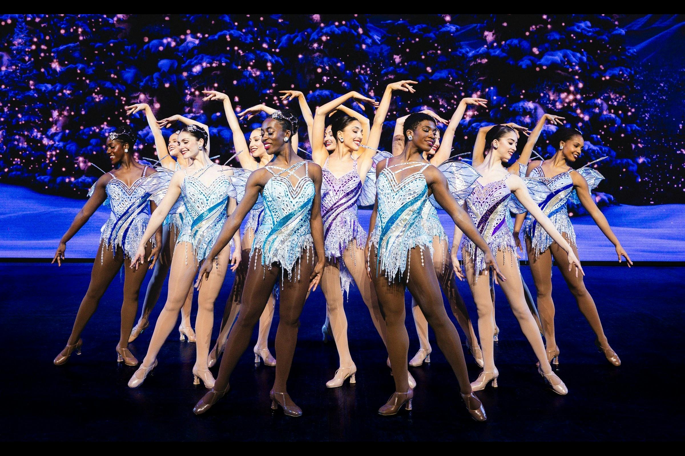 A group of dancers in sparkly costumes performing on a stage with a backdrop of a snowy, starry scene.