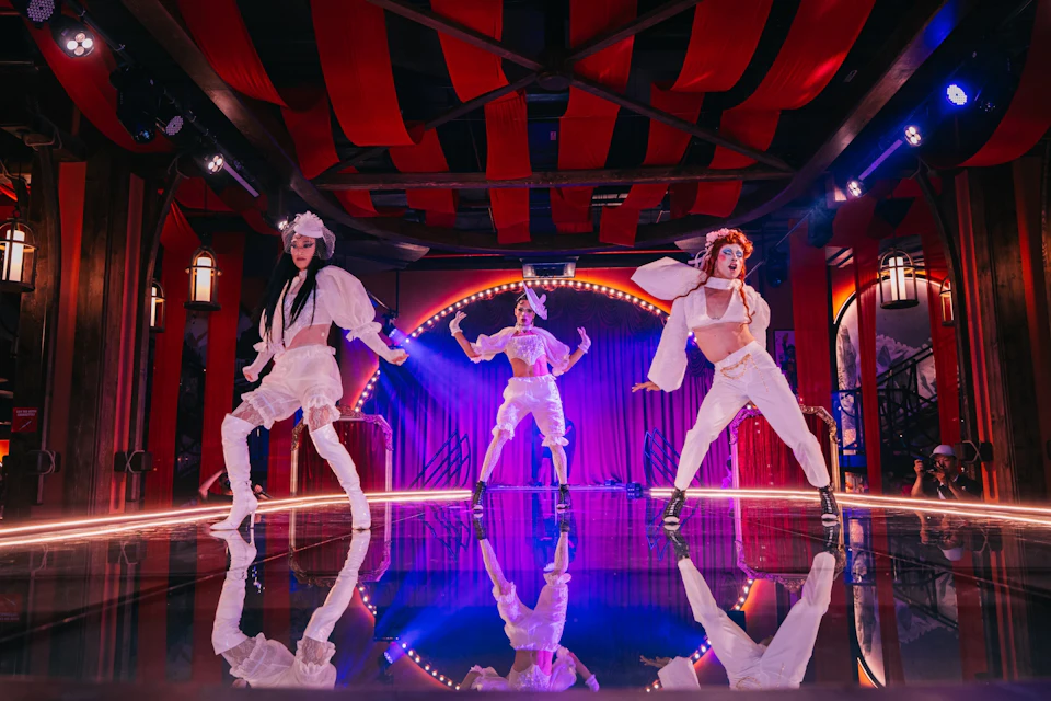 Three performers in extravagant white costumes pose on a lit stage with red and purple decor.
