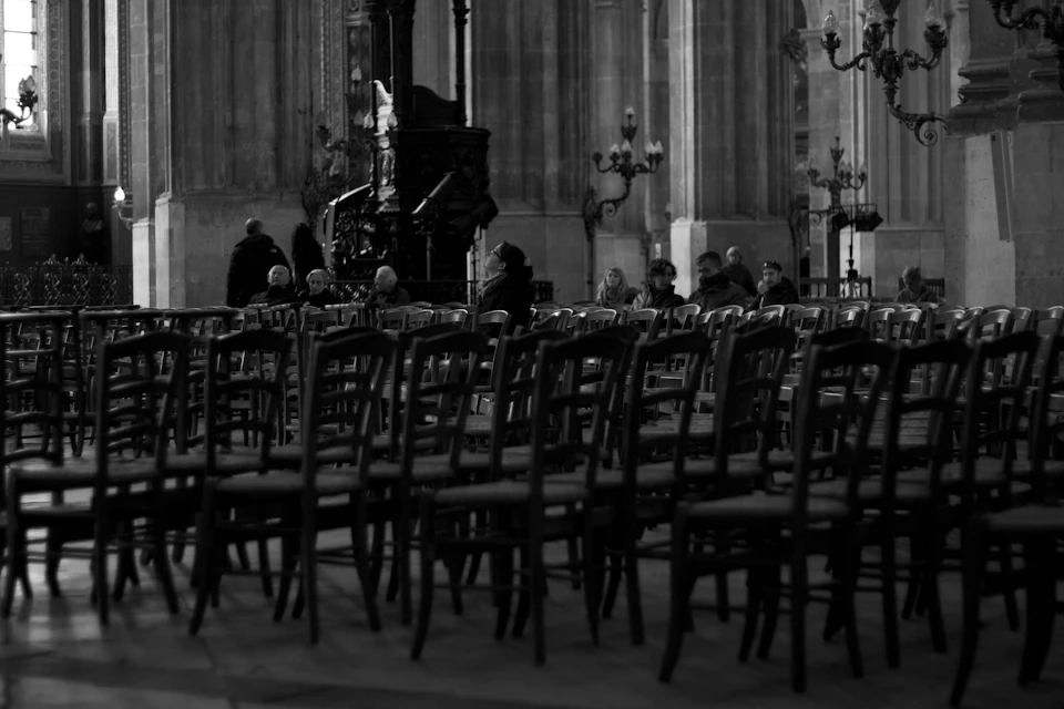 Rows of empty wooden chairs inside a church with few people sitting in the back, and ornate architecture visible in the background.