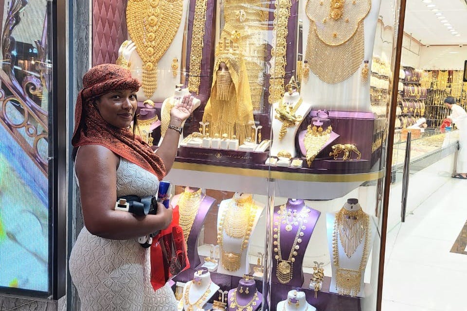 Μια γυναίκα με ελαφρύ φόρεμα και μαντίλι στέκεται μπροστά από μια βιτρίνα καταστήματος παρουσιάζοντας διάφορα χρυσά κοσμήματα.