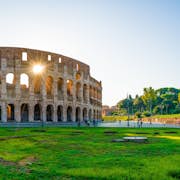 L'antico Colosseo e l'Arco di Costantino a Roma, con erba verde e visitatori sparsi sotto un cielo azzurro e limpido.