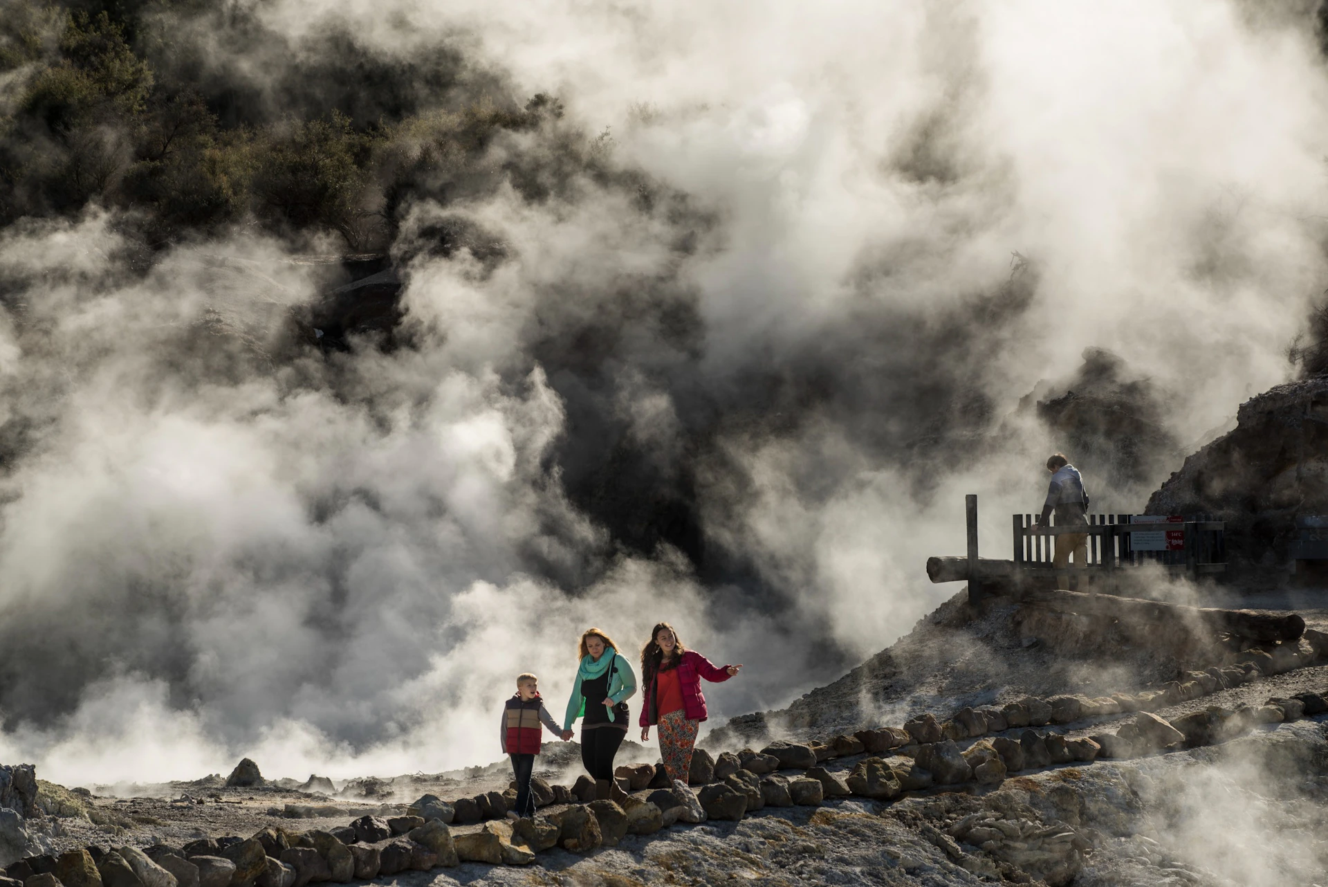Three people walk along a rocky path near a steaming geothermal area, with a wooden railing visible in the background.