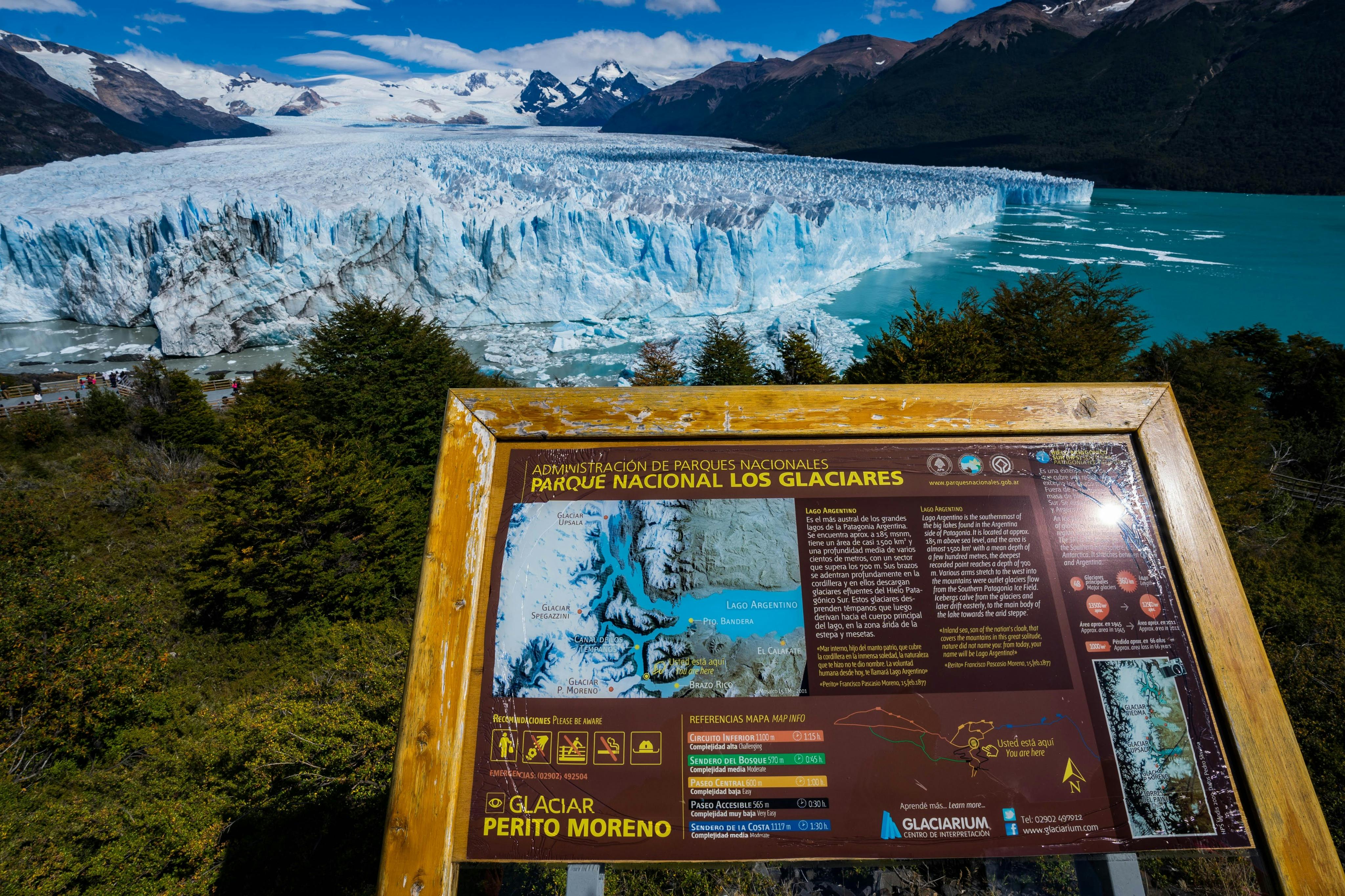 A scenic view of Perito Moreno Glacier with an informational signboard in the foreground, surrounded by vegetation.