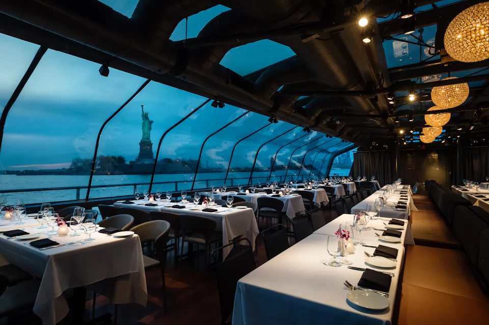 Elegant dining area inside a glass-enclosed boat with set tables and views of the Statue of Liberty outside.