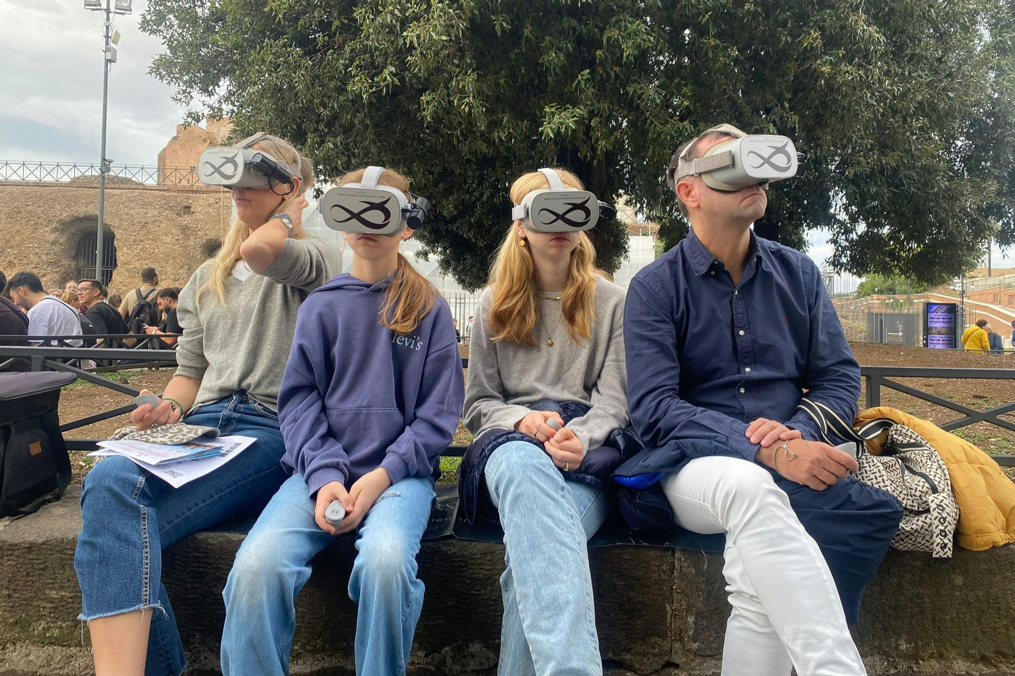 Tre persone sedute su una panchina all’aperto con indosso dei visori per la realtà virtuale, con un sito storico e un albero sullo sfondo.