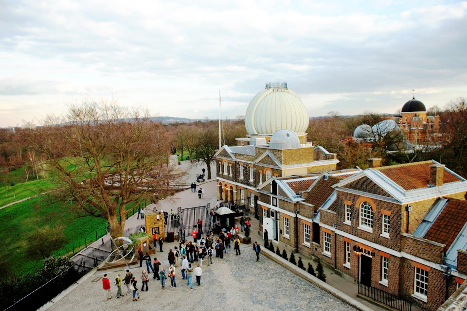 A large group of people stands near an entrance gate and a historic building with a domed observatory, set in a park.