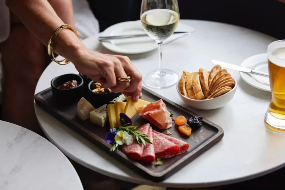 A hand reaches for food on a charcuterie board with meats, cheeses, and fruit. Nearby are crackers and a glass of white wine.