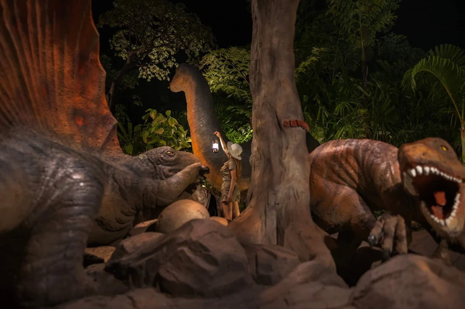 A person in safari attire interacts with life-sized dinosaur statues in a jungle-themed setting at night.
