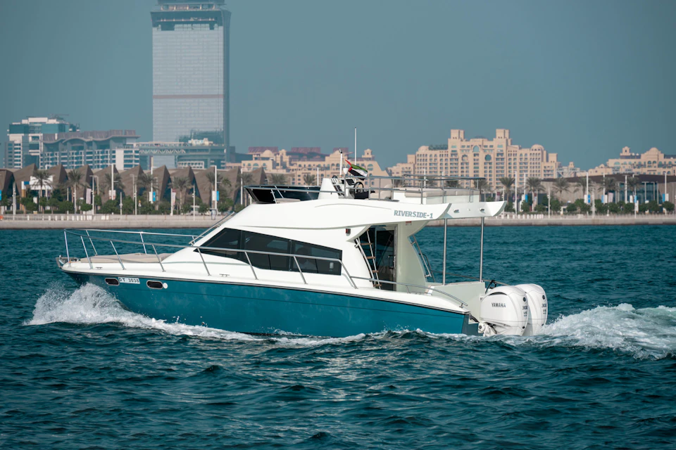 A white and blue motor yacht cruising on the water with a city skyline in the background under a clear sky.