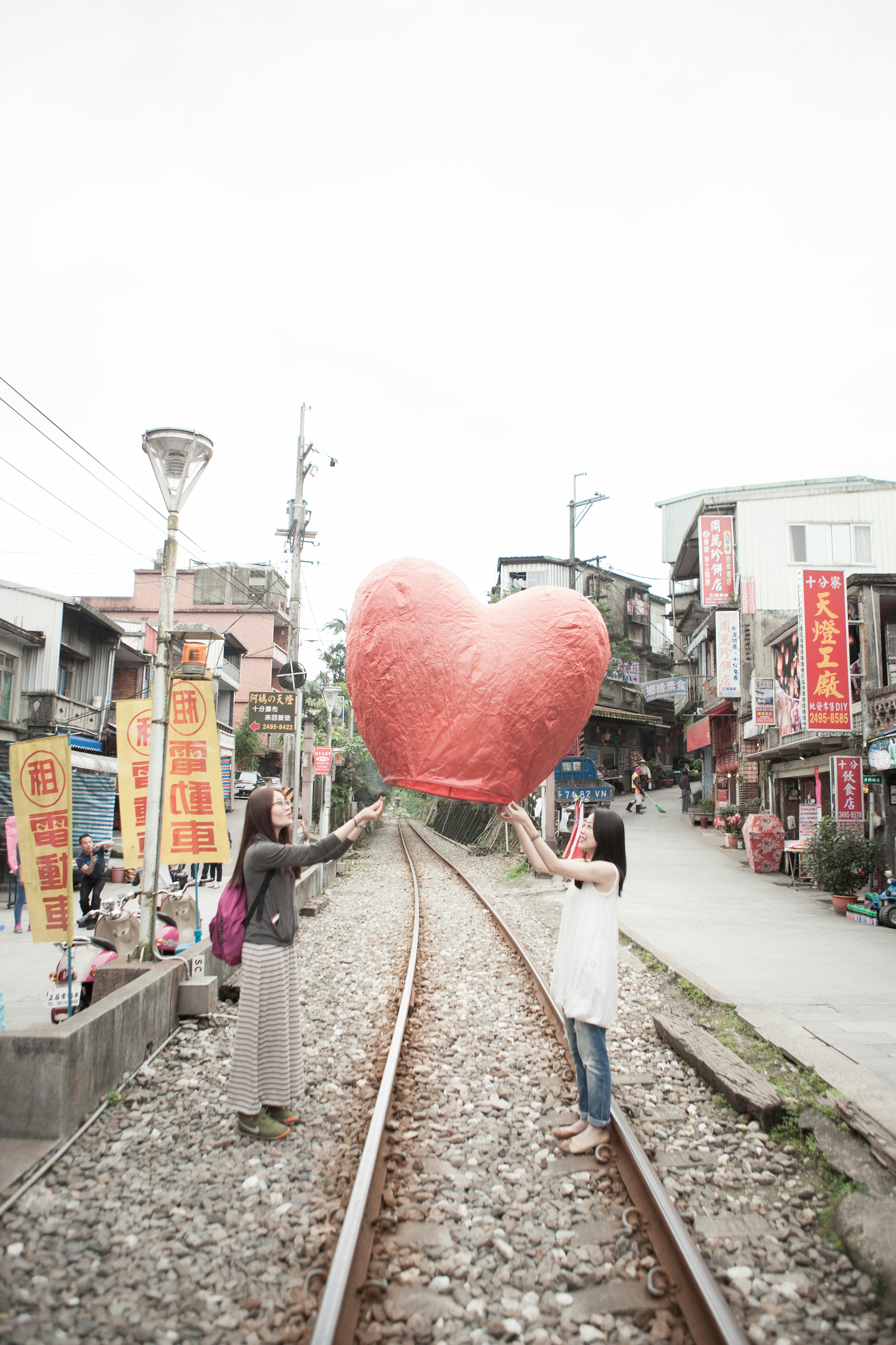2人の女性が、ショップや看板が背景にある都市部の鉄道線路に、大きなハート型のランタンを放ちます。