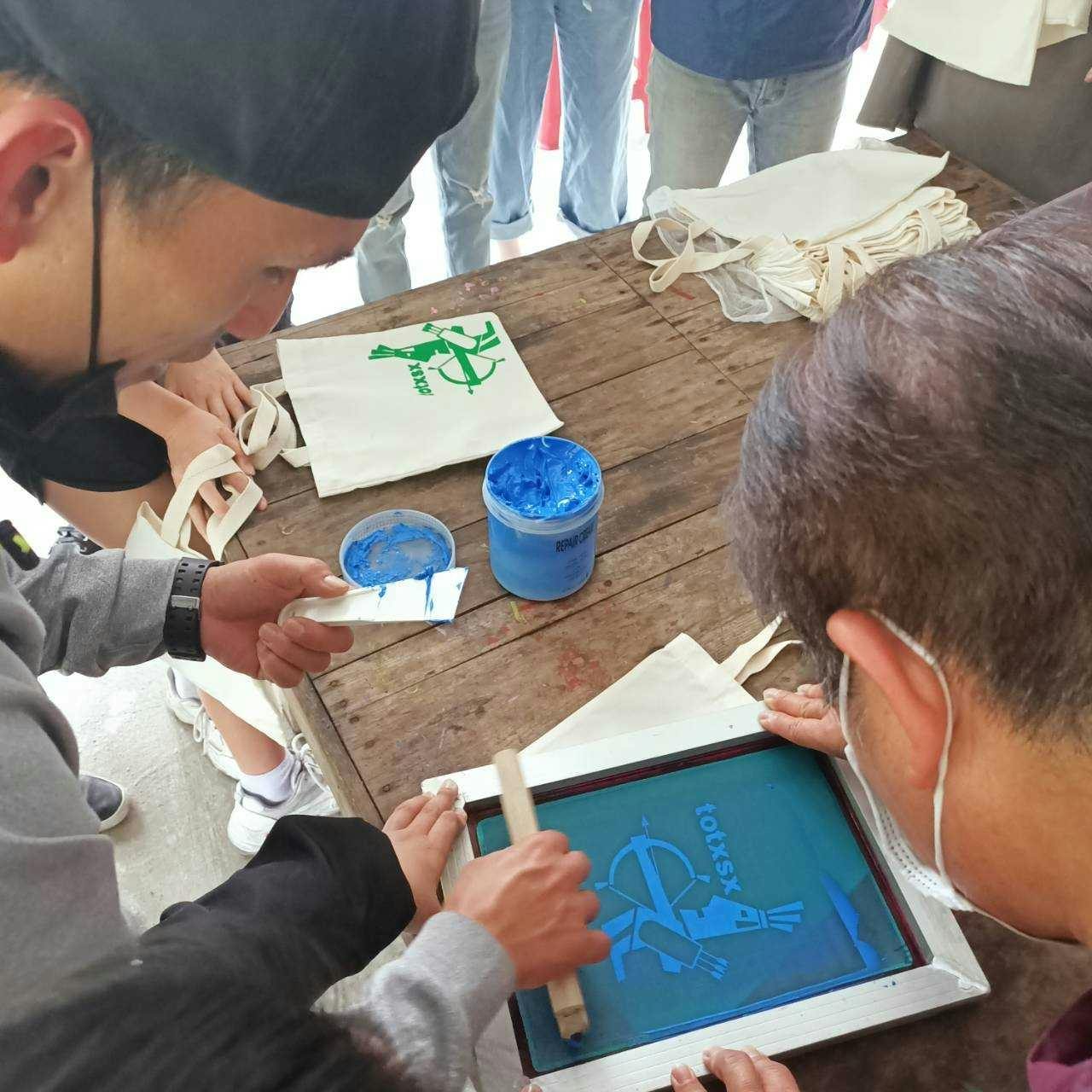 People engaging in screen printing on a wooden table, creating designs on fabric with green and blue paint.