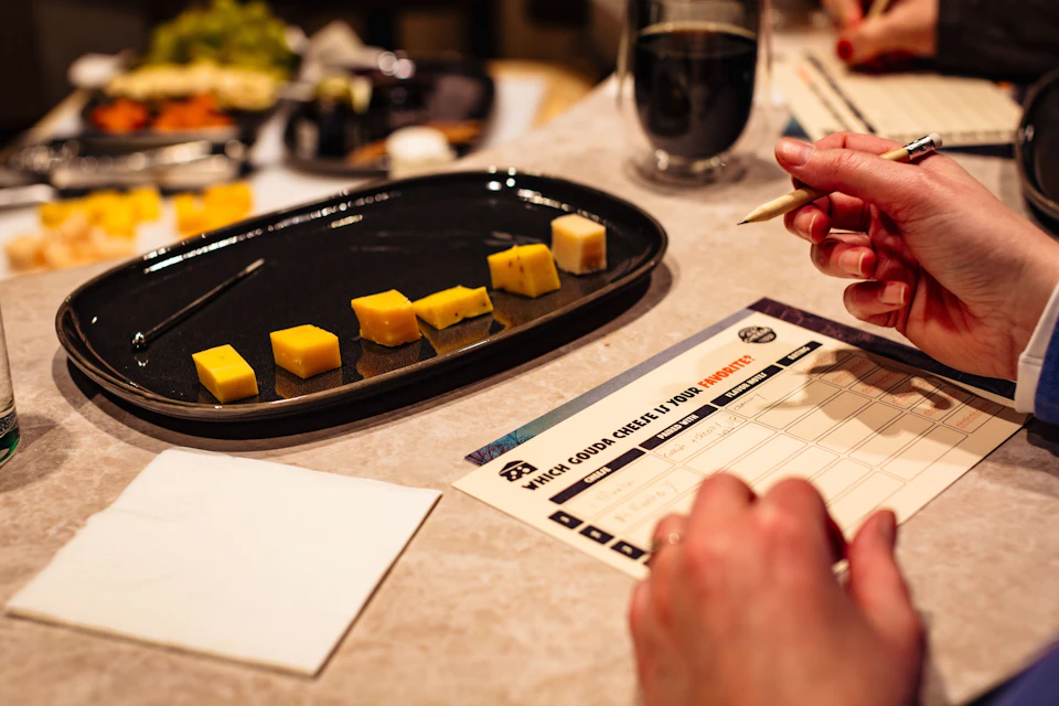 A person holding a pen examines a tasting card next to a black platter with cubes of cheese on a table.