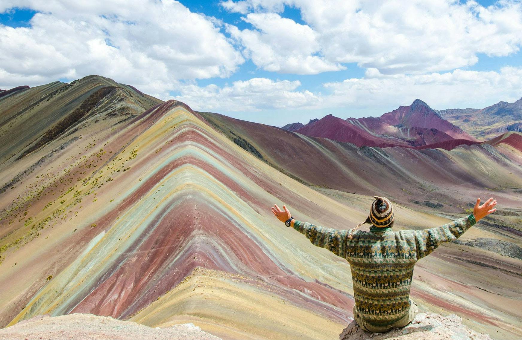 Person with arms outstretched sitting on a rock, overlooking colorful, striped mountains under a partly cloudy sky.