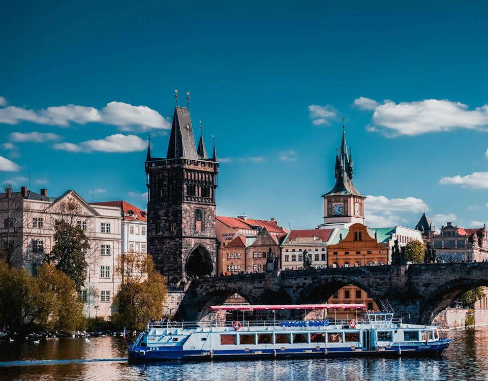 A boat on a river in front of a historic cityscape with a bridge, gothic tower, and clock tower under a blue sky with clouds.