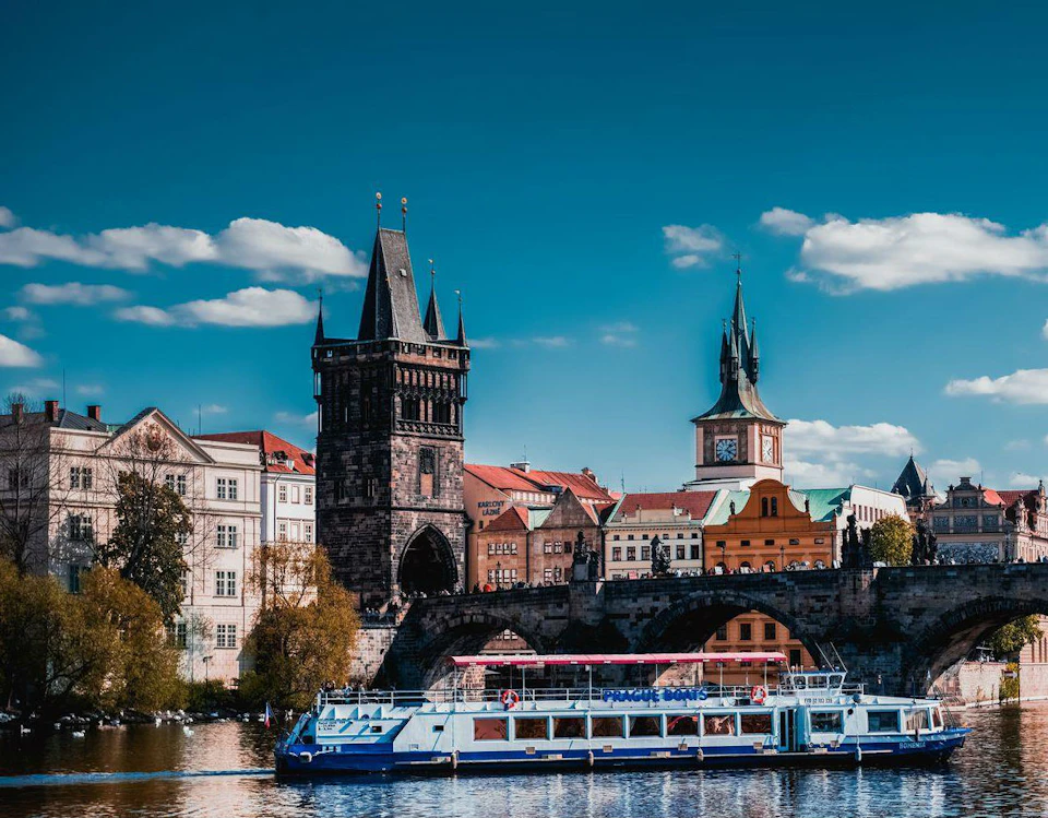 A boat on a river in front of a historic cityscape with a bridge, gothic tower, and clock tower under a blue sky with clouds.