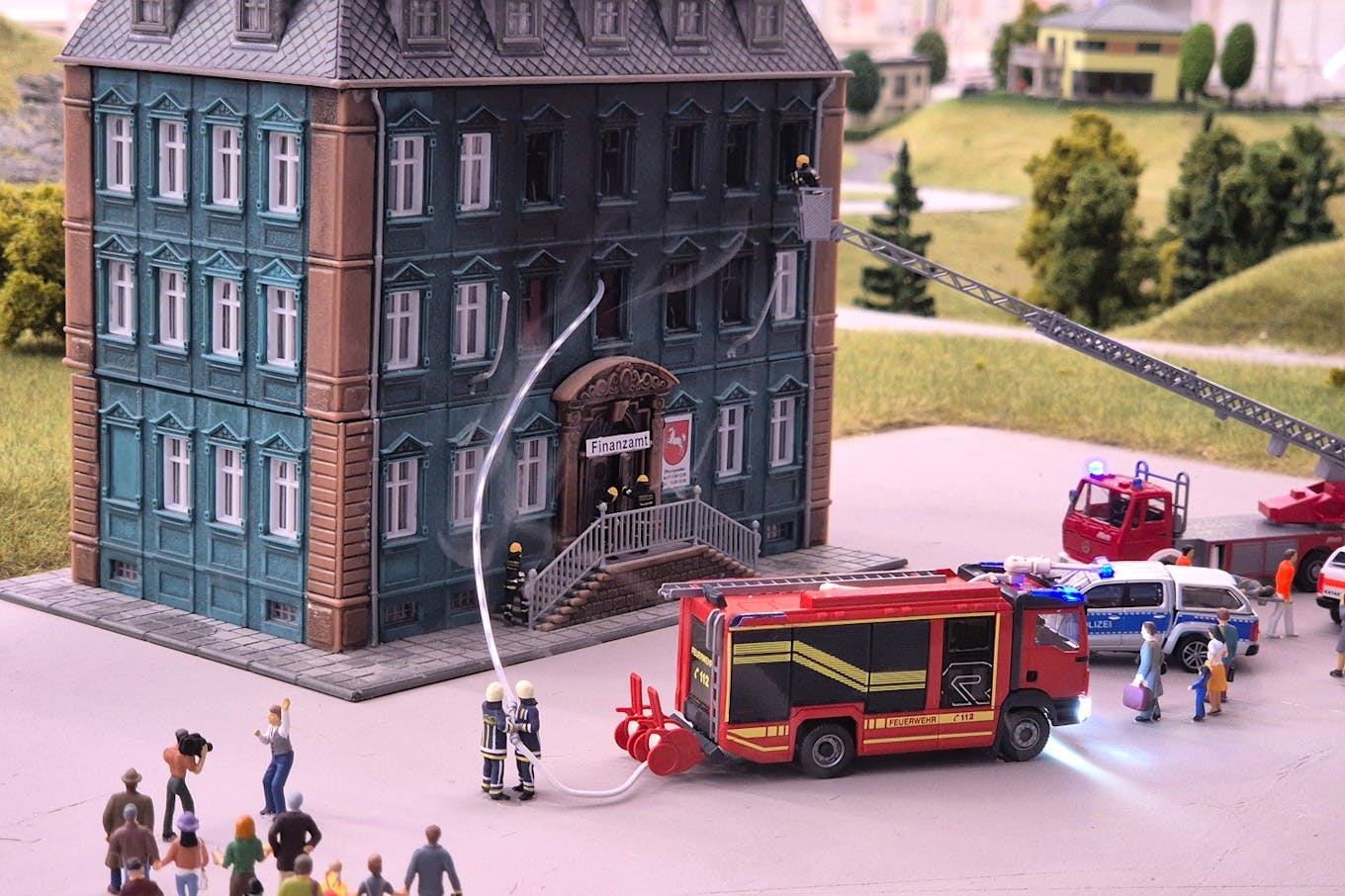 Firefighters use a ladder truck and hose to attend to a building labeled "Finanzamt" as onlookers watch.
